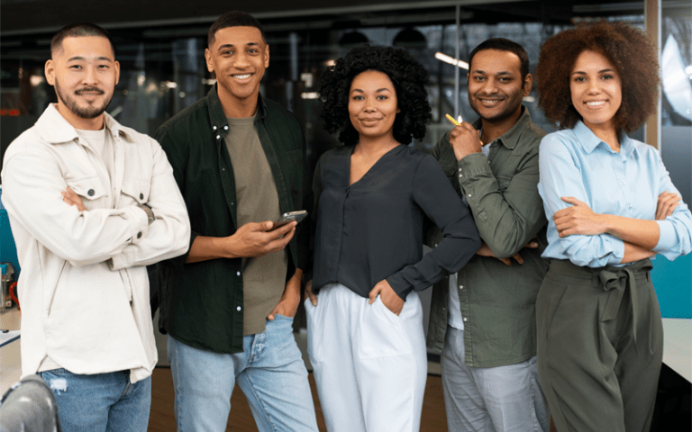Grupo diverso de cinco jovens profissionais sorri lado a lado em um ambiente corporativo moderno. Composto por homens e mulheres de diferentes etnias e traços, o grupo representa a representatividade, a inclusão e o impacto das vagas afirmativas na construção de equipes plurais e inovadoras.