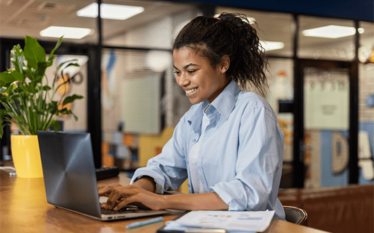 Uma profissional jovem, negra, sorri de forma confiante enquanto trabalha autonomamente em seu notebook. Ela está em um escritório moderno, focada e segura, personificando a autonomia no trabalho.