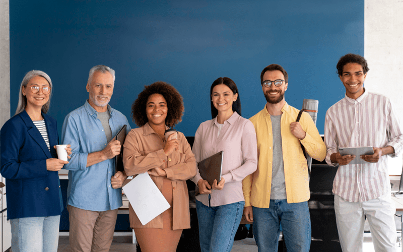Equipe corporativa diversa sorri para a foto em um escritório com parede azul ao fundo. O grupo de seis profissionais, composto por homens e mulheres de diferentes etnias e idades, segura notebooks, tablets e relatórios impressos, representando a diversidade de perfis comportamentais e talentos em uma organização.
