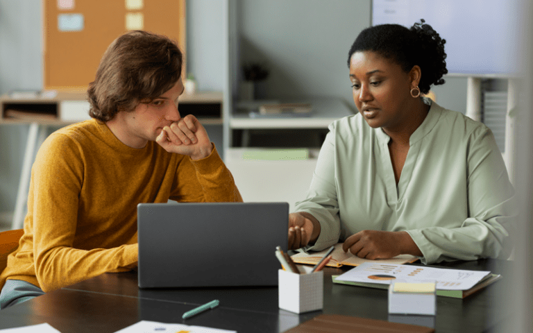 Mulher negra com cabelo preso e blusa verde-clara conversa e aponta para a tela de um laptop, orientando um homem branco de blusa amarela mostarda que a escuta atentamente com a mão no queixo. A imagem simula uma devolutiva comportamental em ambiente corporativo.