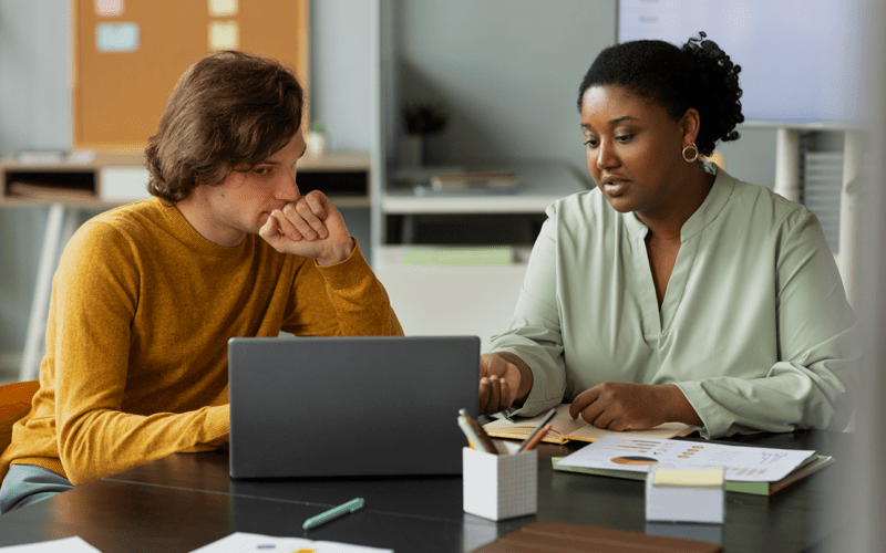 Mulher negra com cabelo preso e blusa verde-clara conversa e aponta para a tela de um laptop, orientando um homem branco de blusa amarela mostarda que a escuta atentamente com a mão no queixo. A imagem simula uma devolutiva comportamental em ambiente corporativo.