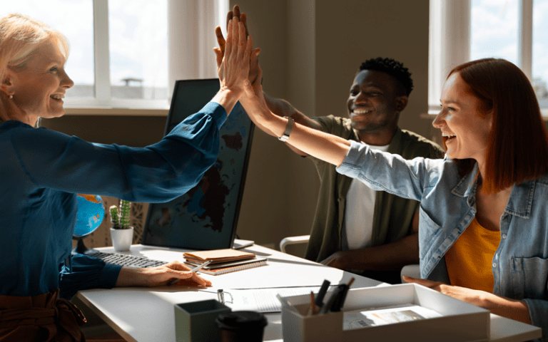 Uma líder e sua equipe celebram uma conquista sobre uma mesa de escritório. A líder (mulher loira à esquerda) e dois colaboradores (homem negro ao fundo e mulher ruiva à direita) sorriem e dão as mãos no ar, demonstrando inteligência emocional em um ambiente de trabalho acolhedor e produtivo.