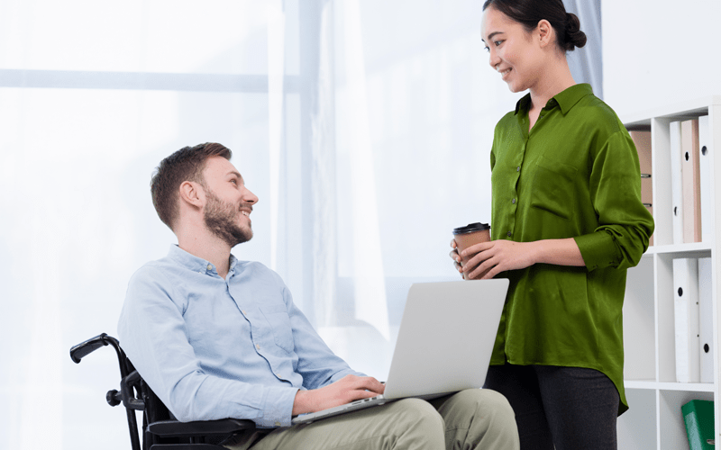 Interação profissional e inclusiva em escritório. Homem cadeirante utiliza notebook e sorri ao conversar com colega de trabalho que está de pé ao seu lado. Ambos demonstram sintonia e colaboração em um ambiente de trabalho acessível.