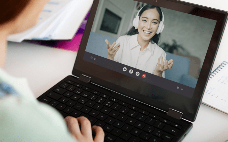 Visão em primeira pessoa sobre o ombro de uma profissional interagindo em uma chamada de vídeo no laptop com uma colega sorridente que usa fones de ouvido. A tela exibe a interface da videoconferência. A imagem ilustra a importância da liderança digital para as empresas.