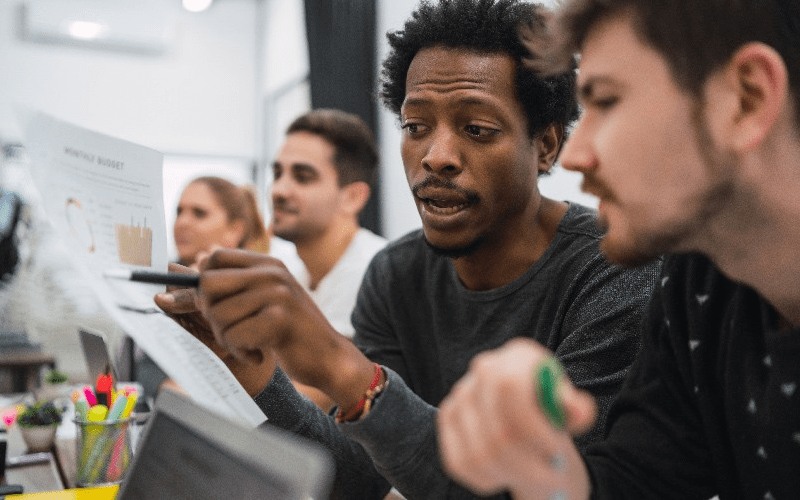 Imagem simula uma equipe de Recursos Humanos realizando análise preditiva de dados. Um profissional negro foca a atenção em um relatório impresso com gráficos de desempenho, apontando com uma caneta e discutindo as métricas com um colega de trabalho em um escritório.