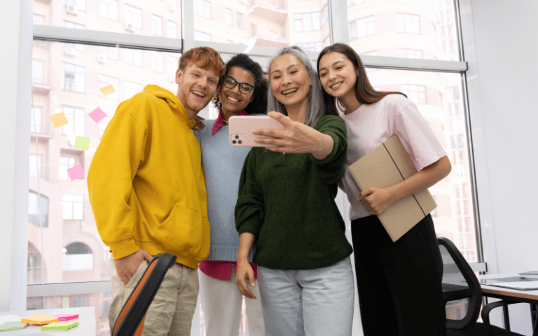 Quatro profissionais de diferentes idades e etnias sorriem juntos para uma selfie tirada com um smartphone em um escritório. Uma mulher madura segura o celular à frente de três jovens colaboradores da Geração Z, ilustrando a integração multigeracional no trabalho.