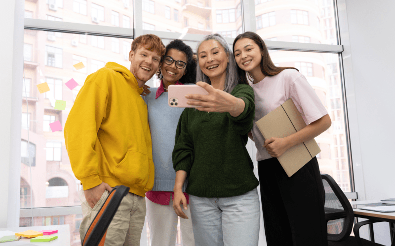 Quatro profissionais de diferentes idades e etnias sorriem juntos para uma selfie tirada com um smartphone em um escritório. Uma mulher madura segura o celular à frente de três jovens colaboradores da Geração Z, ilustrando a integração multigeracional no trabalho.