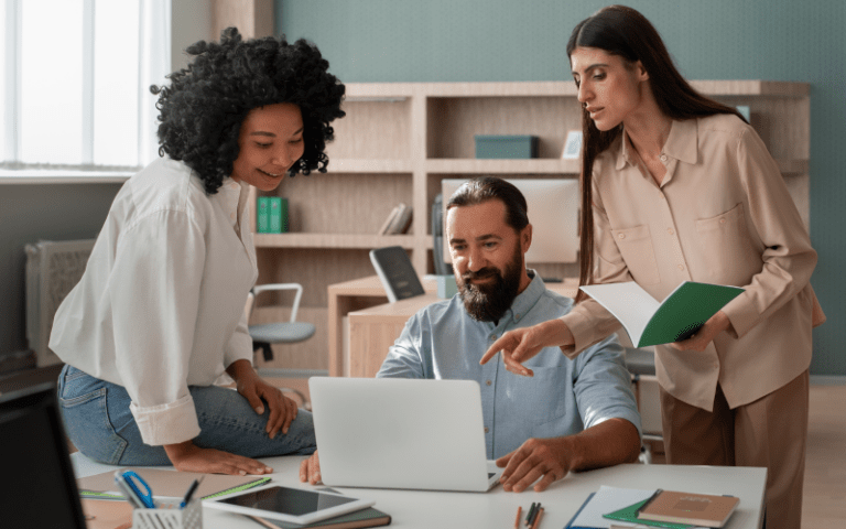 Três profissionais de uma equipe de RH analisam dados juntos em um notebook, trabalhando de forma colaborativa. Uma mulher em pé aponta para a tela enquanto um homem e uma mulher negra observam, ilustrando a visão global do RH sistêmico.