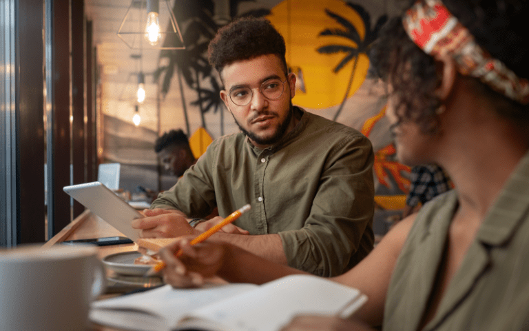 Jovem segurando um tablet e conversando com colegas em ambiente de trabalho descontraído, com caderno de anotações e xícara de café na mesa, representando um momento de diálogo sobre direitos e deveres no estágio.
