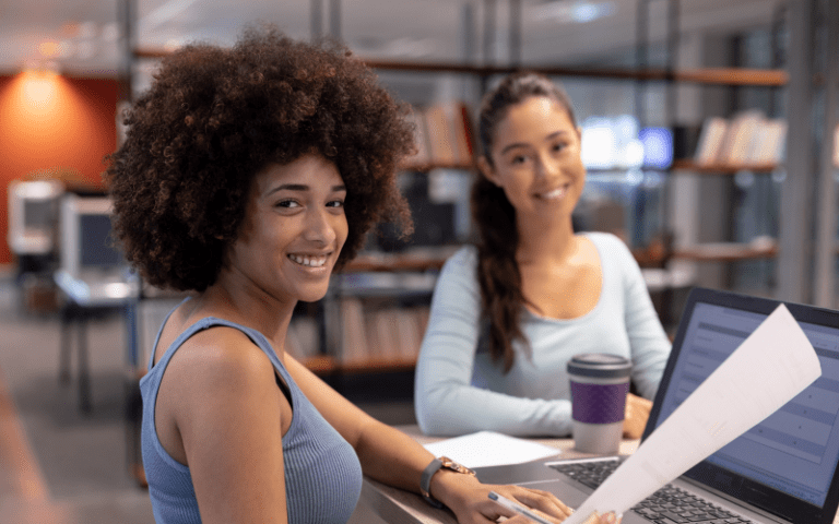 Duas jovens profissionais sorrindo em ambiente de escritório, uma segurando documentos e outra em frente a um notebook, representando um momento de treinamento e integração de estagiários.