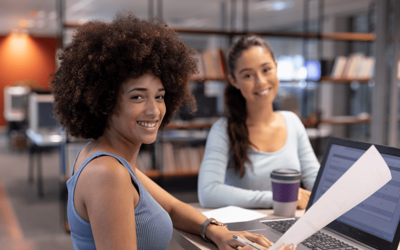 Duas jovens profissionais sorrindo em ambiente de escritório, uma segurando documentos e outra em frente a um notebook, representando um momento de treinamento e integração de estagiários.