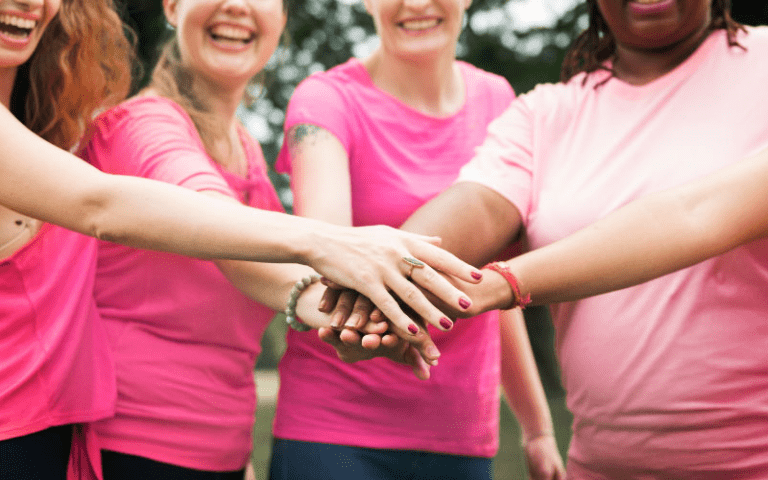 Imagem de capa de artigo sobre outubro rosa nas empresas, mostrando um grupo de pessoas sorrindo e unindo as mãos no centro, criando um momento de apoio e união ao ar livre, com roupas em tons de rosa.magem de capa de artigo sobre outubro rosa nas empresas, mostrando um grupo de pessoas sorrindo e unindo as mãos no centro, criando um momento de apoio e união ao ar livre, com roupas em tons de rosa.