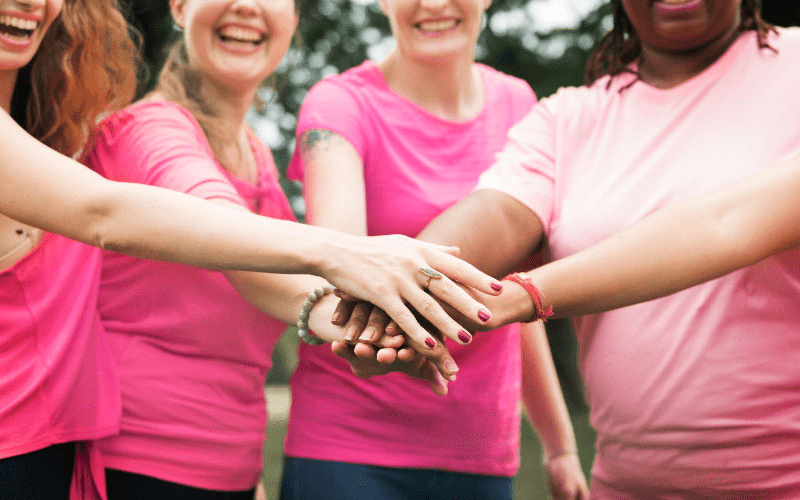 Imagem de capa de artigo sobre outubro rosa nas empresas, mostrando um grupo de pessoas sorrindo e unindo as mãos no centro, criando um momento de apoio e união ao ar livre, com roupas em tons de rosa.magem de capa de artigo sobre outubro rosa nas empresas, mostrando um grupo de pessoas sorrindo e unindo as mãos no centro, criando um momento de apoio e união ao ar livre, com roupas em tons de rosa.