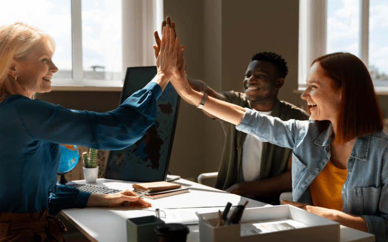 Imagem de capa para artigo sobre Empowerment. Uma gestora loira e sua equipe, composta por um homem negro e uma mulher ruiva, sorriem entusiasmados enquanto comemoram com um "high five" (toque de mãos no alto) sobre a mesa de escritório.