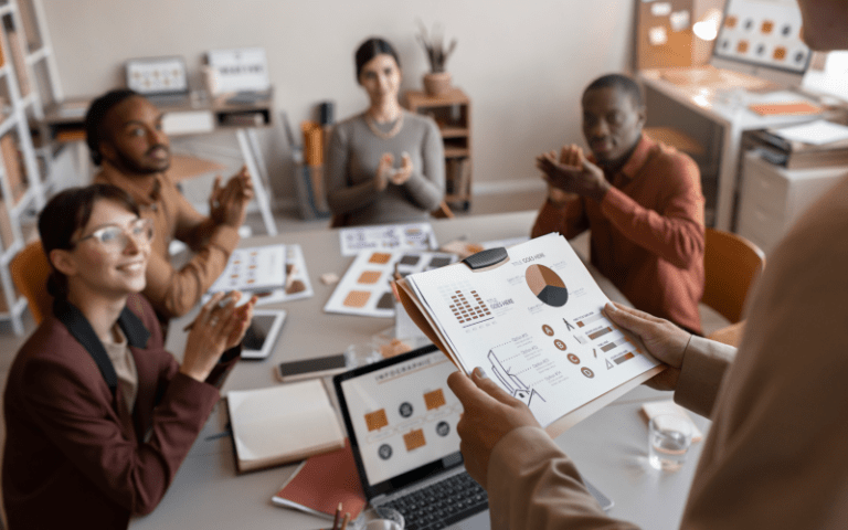 Visão simula líderes do futuro apresentando relatórios com gráficos e infográficos para sua equipe. Ao redor da mesa de reunião, um time diverso de profissionais observa a apresentação com sorrisos e aplausos, demonstrando engajamento e liderança inspiradora.