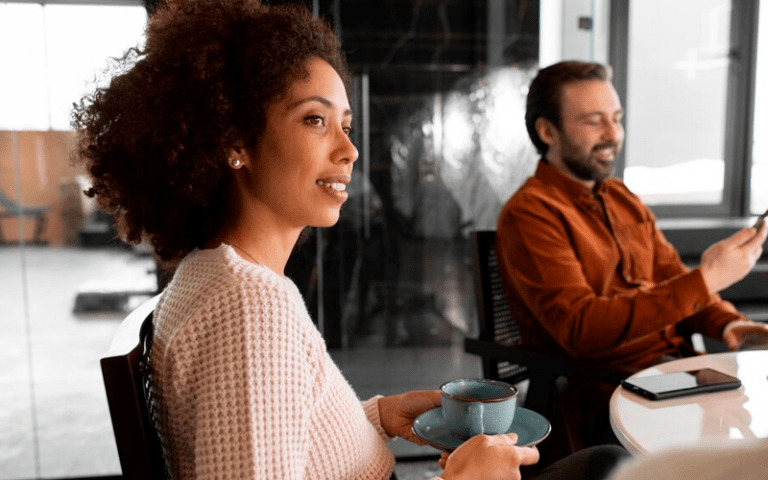 Imagem de capa para artigo sobre Líderes Millennials. Uma jovem mulher negra com cabelo crespo sorri em perfil, segurando uma xícara de café durante uma reunião informal. Ao fundo, um colega de trabalho barbudo mexe no celular em um escritório moderno.