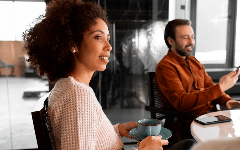 Imagem de capa para artigo sobre Líderes Millennials. Uma jovem mulher negra com cabelo crespo sorri em perfil, segurando uma xícara de café durante uma reunião informal. Ao fundo, um colega de trabalho barbudo mexe no celular em um escritório moderno.