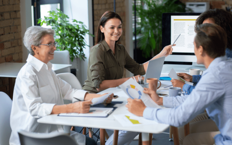 Equipe diversa trabalhando em conjunto em uma mesa de escritório. Uma mulher no centro sorri e aponta para a tela de um computador, facilitando a reunião, enquanto colegas de diferentes idades analisam gráficos impressos e debatem ideias, ilustrando a liderança colaborativa.