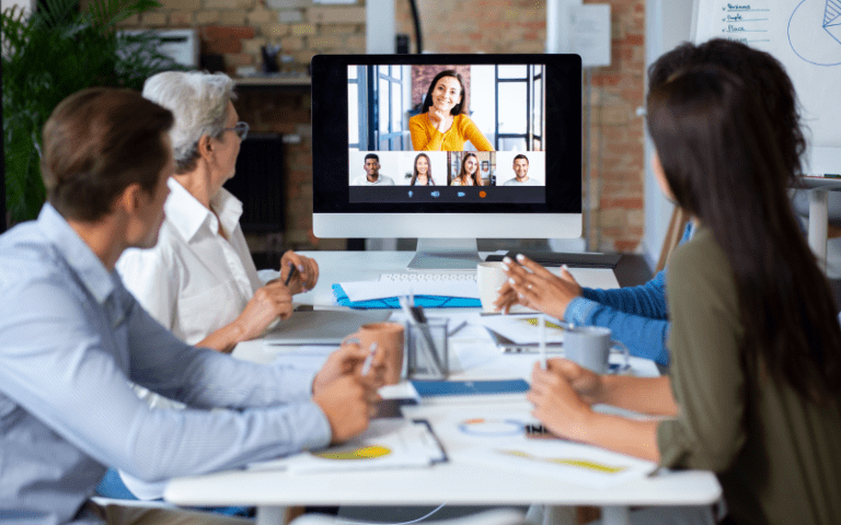 Imagem de capa para artigo sobre universidade corporativa. Grupo de colaboradores reunido em uma mesa de escritório acompanha uma apresentação ou aula online transmitida em um grande monitor de computador.
