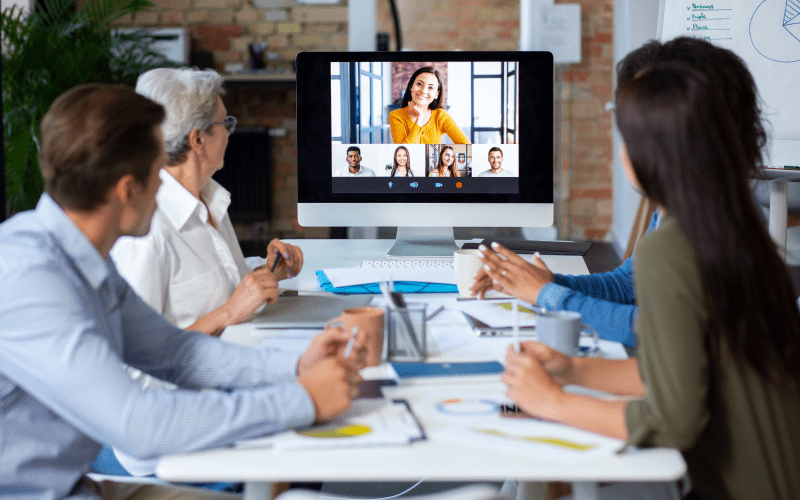 Imagem de capa para artigo sobre universidade corporativa. Grupo de colaboradores reunido em uma mesa de escritório acompanha uma apresentação ou aula online transmitida em um grande monitor de computador.