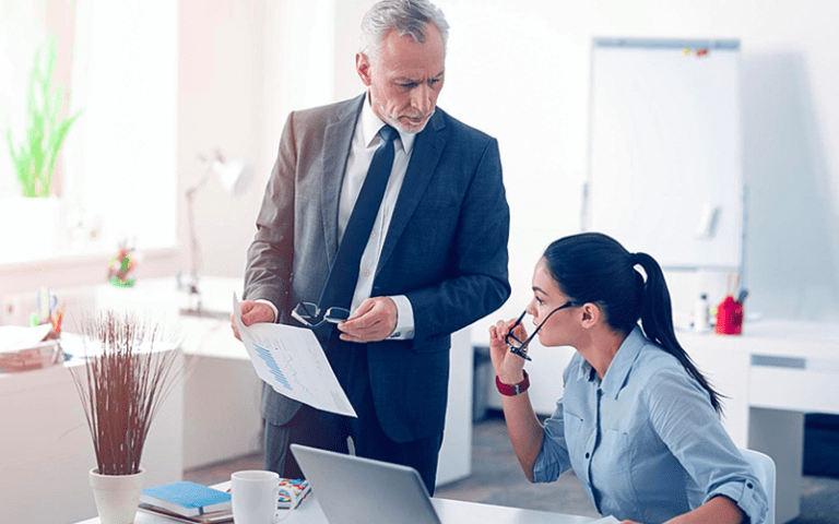 Um homem de terno simulando um líder microgerenciador conversa com sua funcionária sobre uma demanda. O cenário representa um ambiente corporativo, com mesa de trabalho, laptop e quadro branco ao fundo.
