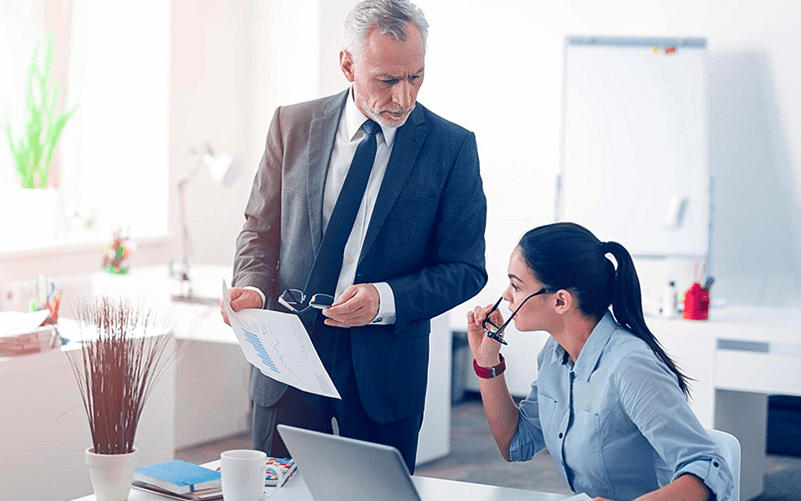 Um homem de terno simulando um líder microgerenciador conversa com sua funcionária sobre uma demanda. O cenário representa um ambiente corporativo, com mesa de trabalho, laptop e quadro branco ao fundo.