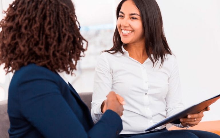 Imagem simula recrutadora e candidata sorrindo enquanto trocam um aperto de mãos acolhedor durante uma entrevista de emprego, simbolizando os pilares de empatia, respeito e conexão do recrutamento humanizado.