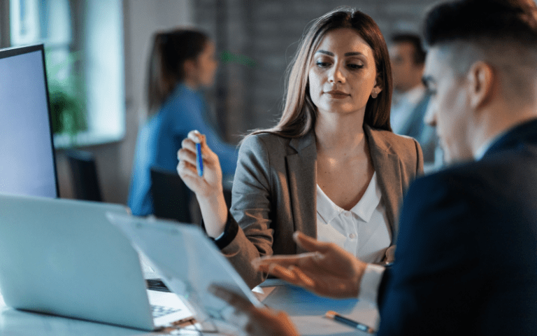 Imagem de capa para artigo sobre Due Diligence. Uma executiva analisa documentos com atenção ao lado de um colega de trabalho em frente a um notebook no escritório.