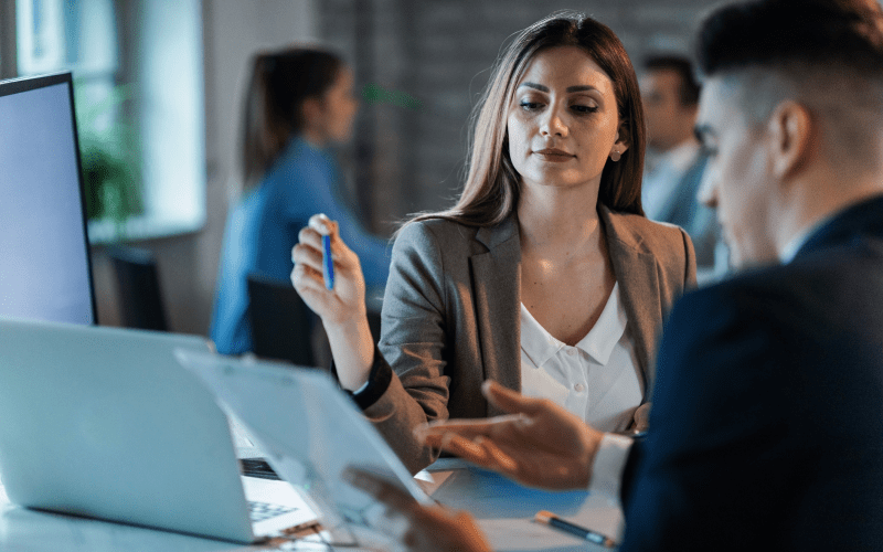 Imagem de capa para artigo sobre Due Diligence. Uma executiva analisa documentos com atenção ao lado de um colega de trabalho em frente a um notebook no escritório.