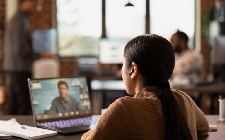 Profissional participa de uma reunião virtual pelo notebook em um ambiente corporativo, enquanto outros colaboradores trabalham ao fundo. A imagem representa o apoio estratégico do RH terceirizado na gestão de pessoas e processos.