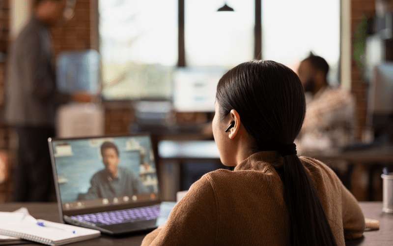 Profissional participa de uma reunião virtual pelo notebook em um ambiente corporativo, enquanto outros colaboradores trabalham ao fundo. A imagem representa o apoio estratégico do RH terceirizado na gestão de pessoas e processos.