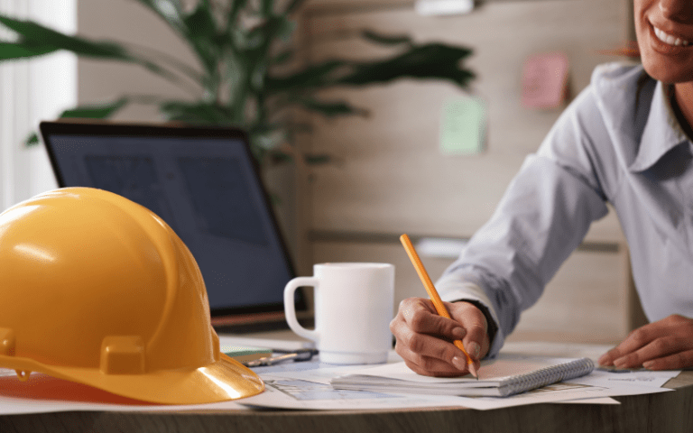 Profissional faz anotações sobre documentos técnicos em uma mesa de trabalho, ao lado de um capacete de segurança e um notebook, ilustrando o planejamento de atividades para atender à demanda complementar de serviços.