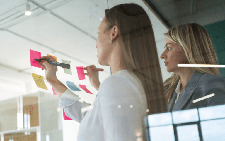 Duas profissionais analisam post-its colados em uma parede de vidro durante uma atividade de planejamento. A cena representa um momento de troca e organização de ideias, simbolizando a importância da análise estruturada de hard skills durante processos seletivos e gestão de equipes.