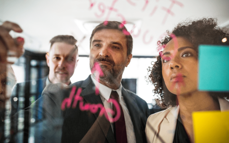 Imagem de capa para artigo sobre consultoria estratégica. Uma mulher negra e dois homens de terno observam atentamente um painel de vidro com gráficos e notas adesivas coloridas.