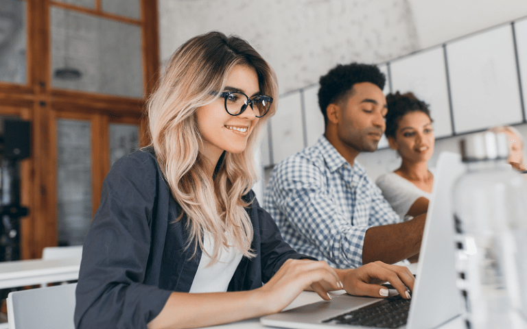 Jovens profissionais em um ambiente corporativo, colaborando em grupo e usando notebooks, representando o perfil de talentos que empresas buscam ao contratar uma consultoria especializada em recrutamento de trainees.