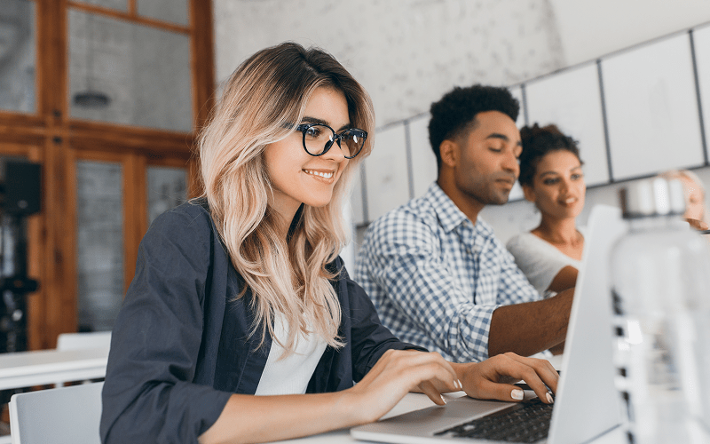 Jovens profissionais em um ambiente corporativo, colaborando em grupo e usando notebooks, representando o perfil de talentos que empresas buscam ao contratar uma consultoria especializada em recrutamento de trainees.