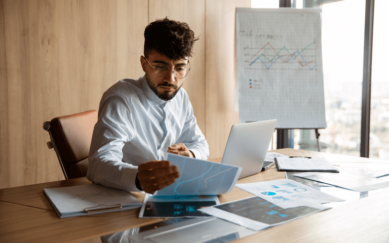 Homem com óculos e camisa social analisando relatórios e gráficos de desempenho sobre uma mesa de escritório, com um laptop e um flipchart ao fundo, sugerindo a aplicação da visão sistêmica nos negócios.