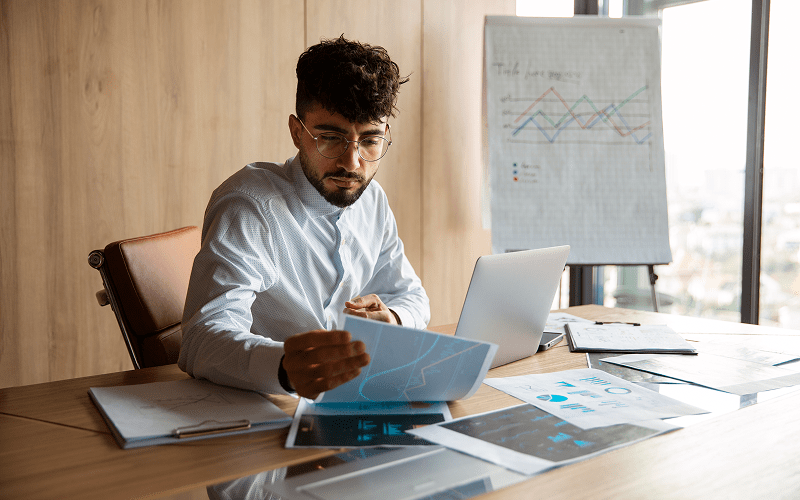 Homem com óculos e camisa social analisando relatórios e gráficos de desempenho sobre uma mesa de escritório, com um laptop e um flipchart ao fundo, sugerindo a aplicação da visão sistêmica nos negócios.