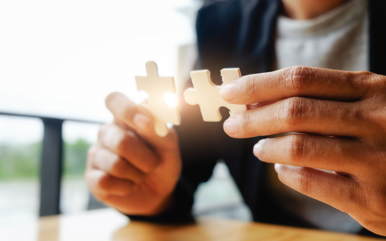 Pessoa encaixando duas peças de um quebra-cabeça, representando a capacidade de resolver problemas, conectar informações e aplicar raciocínio lógico no ambiente corporativo. No recrutamento e seleção, essas habilidades cognitivas são fundamentais para prever performance e identificar o potencial de desenvolvimento das equipes.