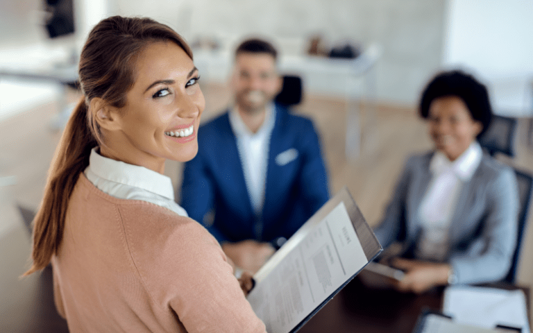 Profissional sorrindo enquanto segura um currículo durante uma entrevista de emprego, com dois recrutadores ao fundo em uma sala de reuniões. A imagem representa um processo seletivo apoiado por decisões estratégicas de recrutamento preditivo.