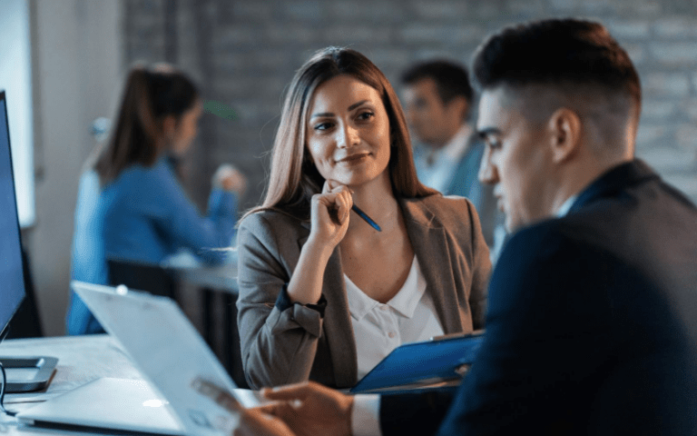 Profissionais em reunião corporativa. Mulher observa atentamente o colega enquanto ele apresenta informações no laptop, transmitindo escuta ativa e perfil de liderança empresarial.