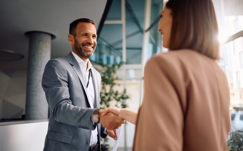 Homem de negócios sorridente aperta a mão de uma mulher em ambiente de escritório, simbolizando sucesso empresarial e networking, importante habilidade de headhunters nas contratações de vagas executivas.