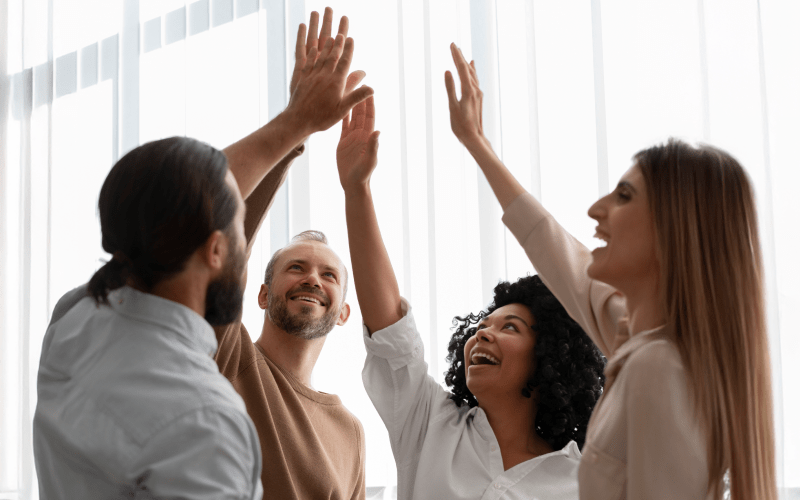 Quatro profissionais celebram juntos com um high-five coletivo, simbolizando união, entusiasmo e o impacto positivo da liderança de alta performance nas equipes.