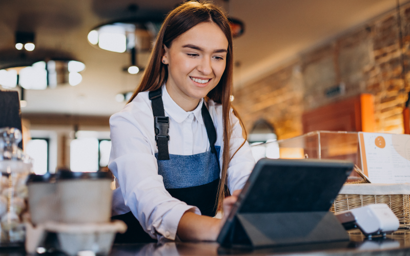 Profissional mulher do varejo usando um tablet no balcão de atendimento, em ambiente comercial, representando a atuação de trabalhadores temporários em períodos de alta demanda.