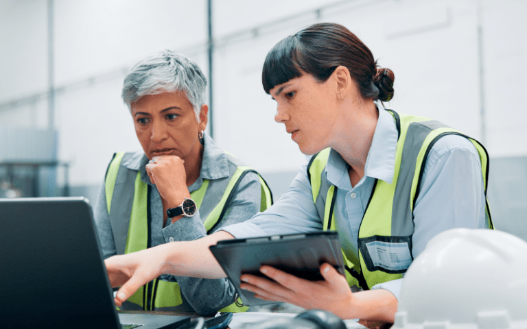 Duas profissionais em ambiente industrial analisam informações em um notebook e um tablet durante uma atividade de acompanhamento de processos, representando práticas de qualidade e gestão no trabalho temporário.